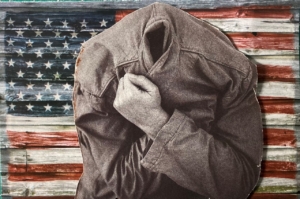 Human with a jacket pulled over their head standing in front of a weathered U.S. flag