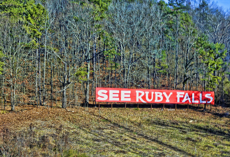 A sign reading See ruby Falls, viewed from across a field with trees in the background.