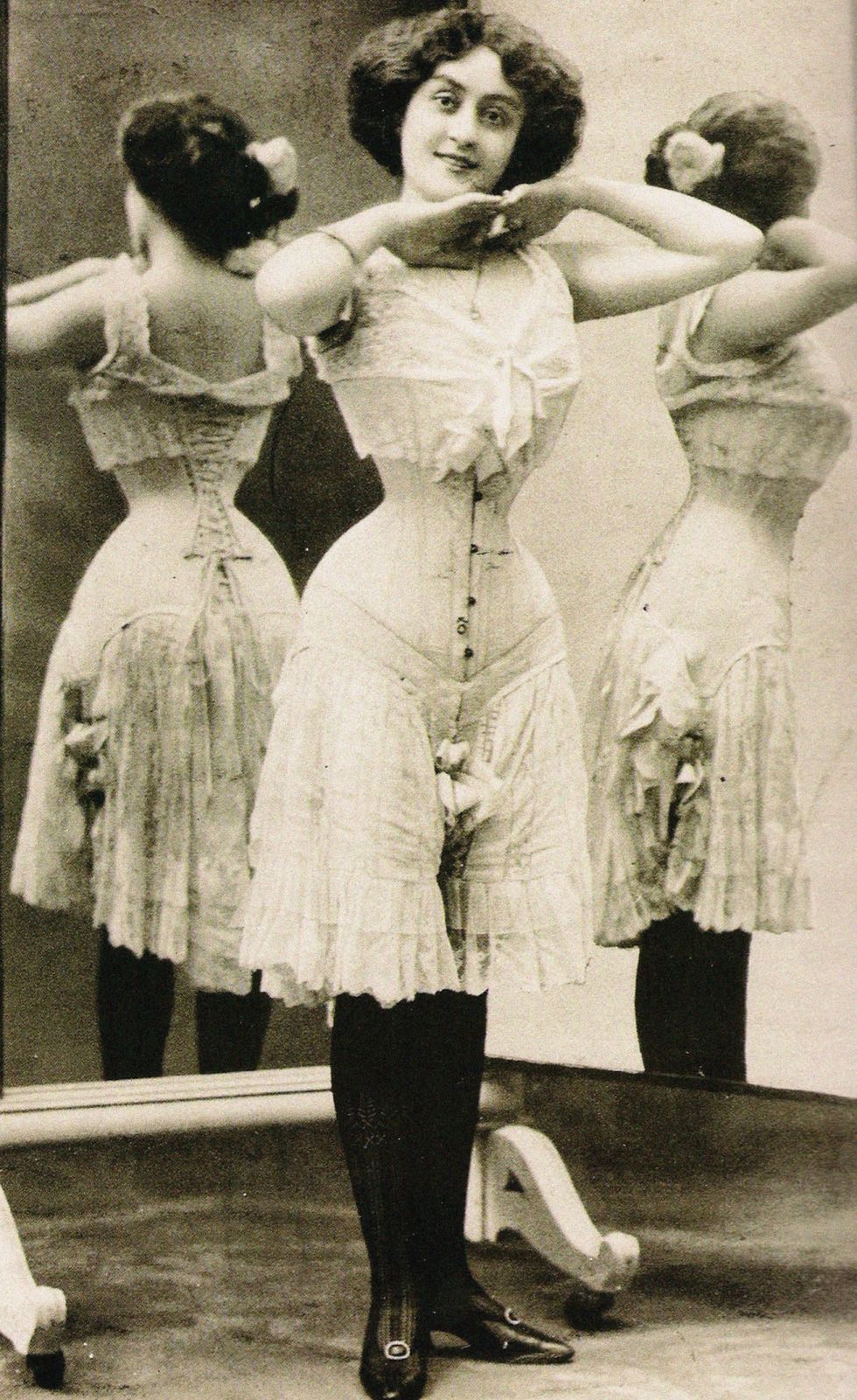 Victorian image of a woman standing before mirrors wearing a corset and bloomers