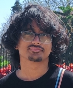 Young person with facial hair, in front of blooming plants and tress