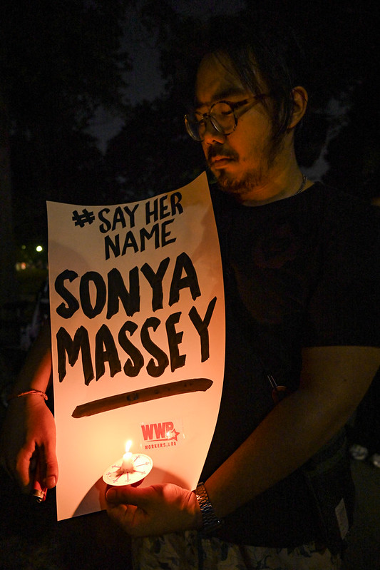 Person at a nighttime vigil, holding a sign reading "Say her name, Sonya Massey."